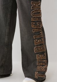 Vaqueros de mezclilla negros con un logotipo marrón bordado texturizado a lo largo del lateral. Corte holgado con costuras estándar y zapatillas blancas visibles en la parte inferior.