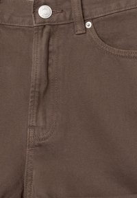 Gros plan de l'avant d'un pantalon marron montrant un bouton métallique, un cache-zipper, une passants de ceinture et une poche droite avec une couture détaillée.