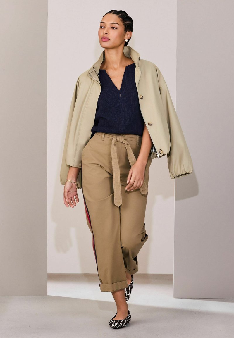 Ženska hodi, oblečena v bež cropped jakno čez mornarsko modro bluzo, svetlo rjave hlače z visokim pasom in stranskimi črtami ter črno-bela vzorčasta balerinka.