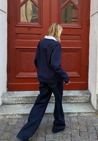 Marineblaues Langarmhemd mit weißem Kragen, kombiniert mit weiten, dunklen Jeans, steht neben einer roten Tür mit rautenförmigen Glasfenstern.