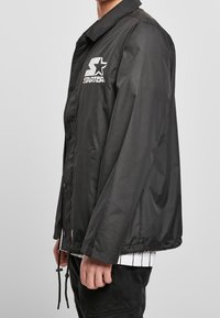 Personne portant une veste Starter noire avec un logo blanc sur la poitrine, devant boutonné et cordons au bas, vue de côté.
