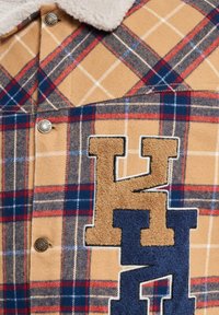Plaid shirt in tan, blue, and red, featuring a shearling collar. Decorative embroidered letters in tan and navy on the front, with metal snaps.