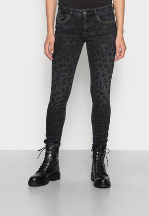 Osoba nosící tmavě šedé skinny džíny s leopardím potiskem a černé šněrovací bojové boty, stojící před jednoduchým světlým pozadím.