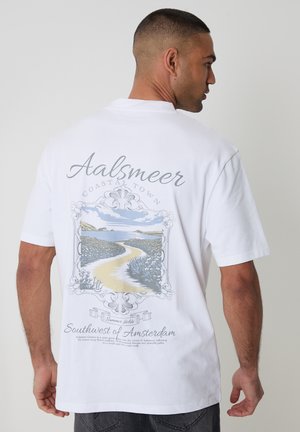 Man draagt een wit T-shirt met een schilderachtig kustlandschap en de tekst "Aalsmeer Coastal Town Southwest of Amsterdam" op de achterkant gedrukt.