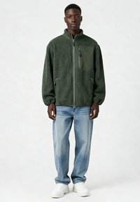 Veste polaire verte avec une fermeture éclair à l'avant, un tissu texturé et une poche plaquée unie. Portée avec un jean bleu clair et des baskets blanches.