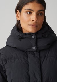 Veste en duvet noire avec un col haut, dotée d'une texture matelassée, de cordons de serrage ajustables et de fermetures à bouton-pression.