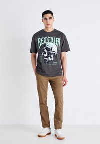 Camiseta gráfica gris con un diseño de calavera y texto verde en negrita, combinada con pantalones beige y zapatillas blancas con detalles azules.