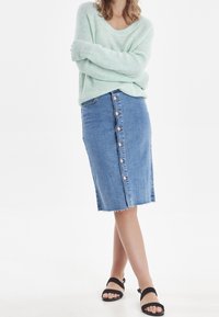 Pull léger menthe, jupe crayon en denim avec boutons à l'avant, ourlet brut, et sandales noires à lanières sur un fond neutre.