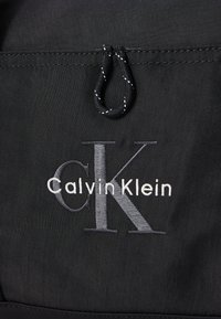 Zwarte stoffen tas met witte en grijze "Calvin Klein" logo-detail. Beschikt over een gestructureerd oppervlak en een trekkoordsluiting.