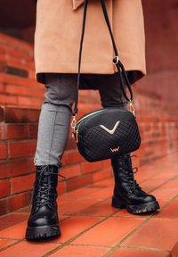 Schwarze gepolsterte Umhängetasche mit goldenen Akzenten und einem grünen Reißverschluss, kombiniert mit schwarzen schnürbaren Kampfstiefeln und grauen, taillierten Hosen.