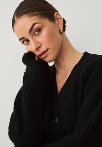 Zwarte cardigan met geribbelde structuur, volumineuze mouwen en een diepe V-halslijn. Voorzien van een enkele zwarte knoopsluiting.