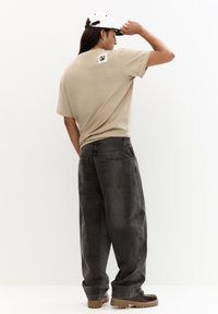 Sandfarvet t-shirt med et firkantet logo patch på ryggen, parret med løstsiddende, mørkegrå jeans og brune fodtøj med strukturerede såler.