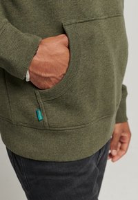 Sudadera verde oliva hecha de una tela suave, con un bolsillo tipo canguro, puños y dobladillo de canalé, y una pequeña etiqueta azul en el lado.