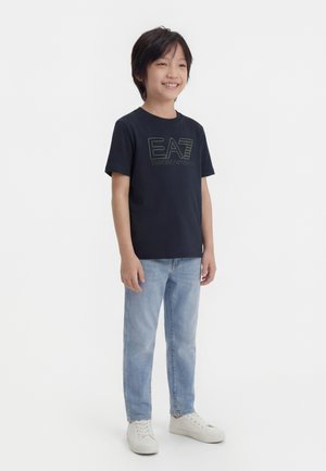 Smiling boy standing, wearing a black EA7 Emporio Armani t-shirt, light blue jeans, and white sneakers against a plain white background.