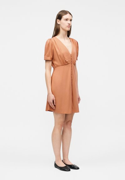 Γυναίκα με μακριά καστανά μαλλιά, που φοράει ένα φόρεμα με κοντά μανίκια, κουμπωτό μπροστά, σε χρώμα σκουριάς, και μαύρες μπαλαρίνες, στέκεται μπροστά σε απλό λευκό φόντο.