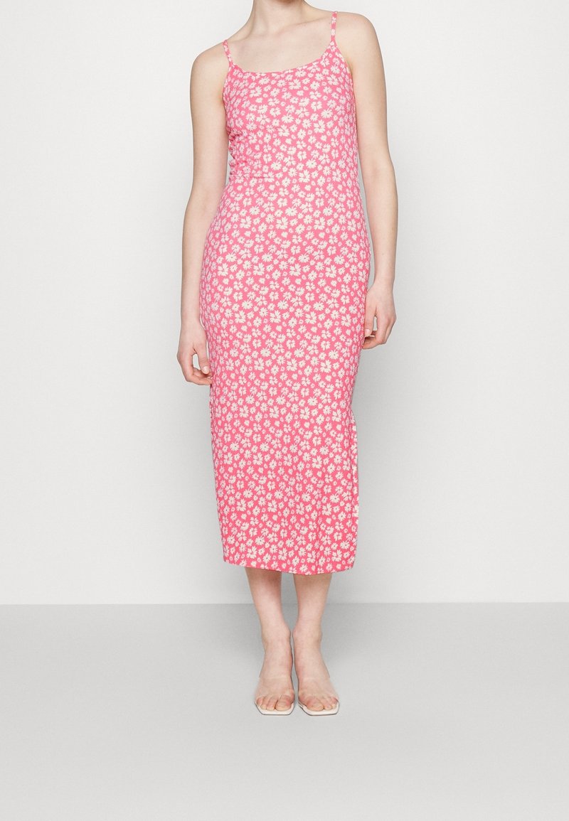 Femme portant une robe rose sans manches, longue jusqu’aux chevilles, à fines bretelles et imprimé floral blanc, debout devant un fond uni.