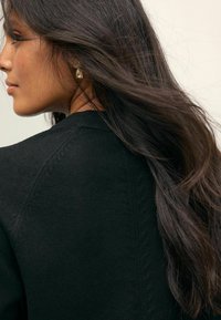 Pull en maille noir avec un col rond, présentant une couture arrière texturée et de longs cheveux châtains foncés flottants en mouvement. Boucles d'oreilles en or visibles.