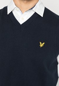 Chaleco de punto azul marino con diseño de cuello en V, que presenta un logo de águila bordado en amarillo en el pecho izquierdo, usado sobre una camisa blanca con cuello.