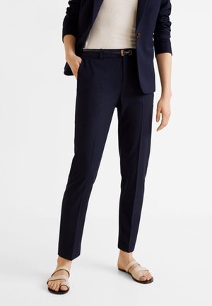 Žena v úzkých tmavomodrých kalhotách, tmavomodrém saku s vyhrnutými rukávy, bílém topu, černém pásku a béžových sandálech stojící na bílém pozadí.