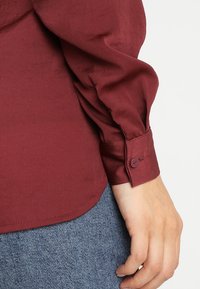 Camicia a maniche lunghe color bordeaux con polsini bottonati. Tessuto liscio e leggero. Orlo arrotondato e lucentezza sottile, abbinata a denim.