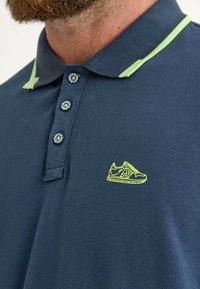 Marinblå pikétröja med en texturerad tyg, gröna krageakcenter, tre knappar och en liten neongul basketsko-broderi på bröstet.