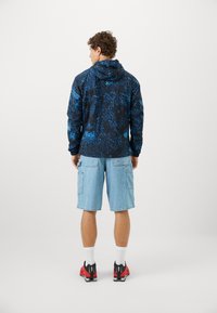 Hættejakke i sort og blå abstrakt mønster, denimshorts og røde sko med sorte detaljer. Korte ærmer og afslappet pasform.