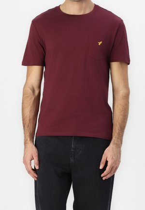 Mand iført en vinrød kortærmet T-shirt med lomme og sorte bukser, stående mod en ensfarvet baggrund.