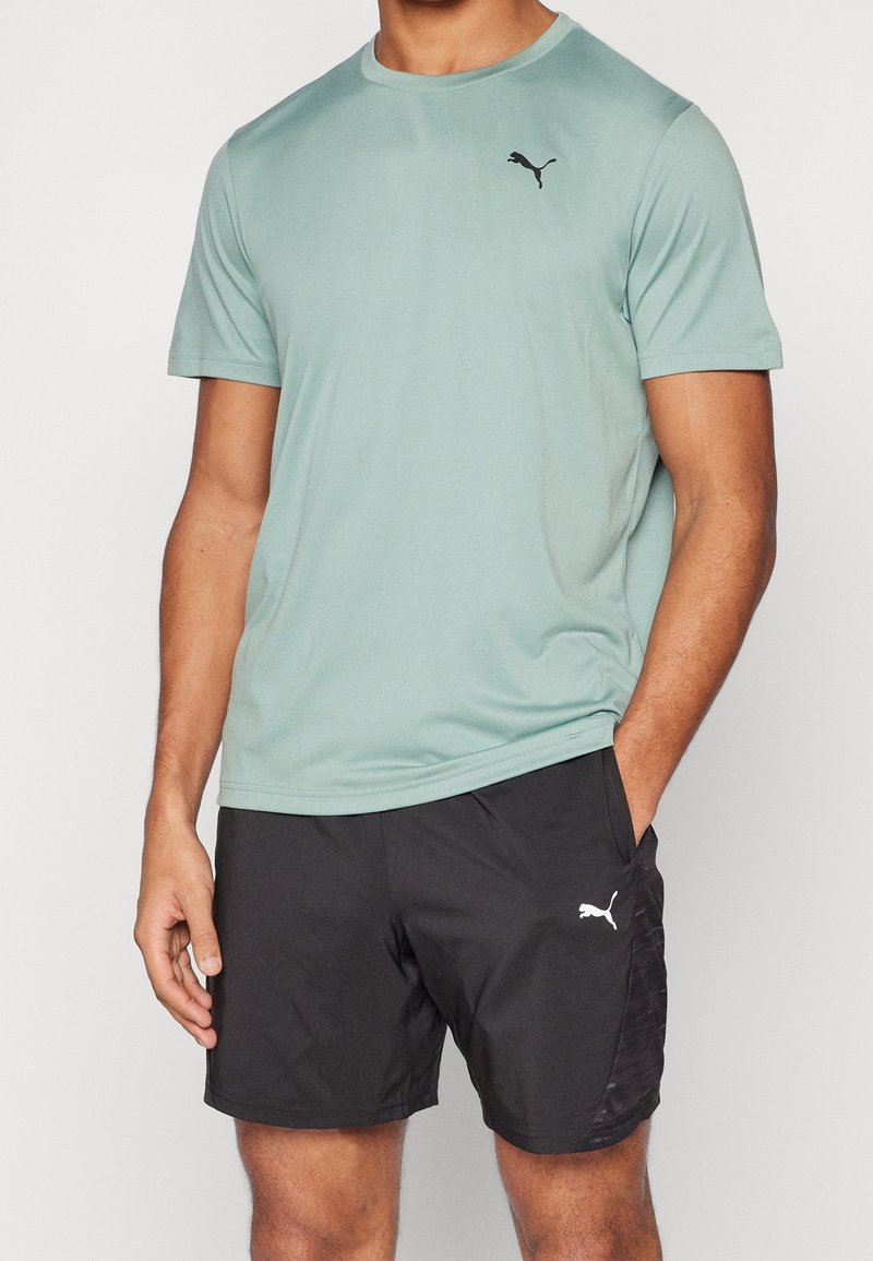 Camiseta deportiva verde claro con cuello redondo, que presenta un logo negro de Puma en el pecho; combinada con pantalones cortos negros de tela texturizada.