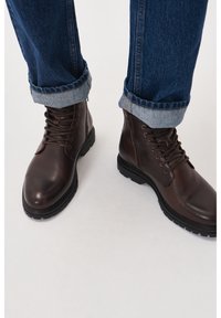 Botas de cuero marrón hasta el tobillo con acabado texturizado, combinadas con suelas de goma oscuras y cordones redondos. Combinadas con unos vaqueros de mezclilla azul arremangados.