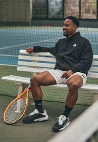 Zwarte jas met een rits, witte shorts met zwarte strepen, oranje tennisracket, zwart-witte sportschoenen, zittend op een witte bank.