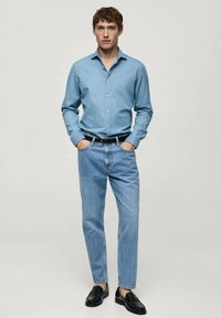 Lichtblauwe overhemd, denimjeans, zwarte riem en zwarte instappers. Het overhemd heeft een kraag en de jeans hebben een relaxte pasvorm.