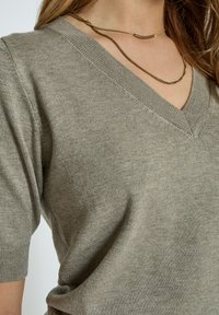 Maglione a collo a V a maniche corte in verde oliva. Caratterizzato da un colletto a costine e una texture liscia con dettagli di cucitura minimi.