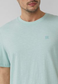 Jasno miętowa koszulka z krótkim rękawem, wykonana z miękkiego, teksturowanego materiału. Posiada małe logo na klatce piersiowej oraz klasyczny okrągły dekolt.