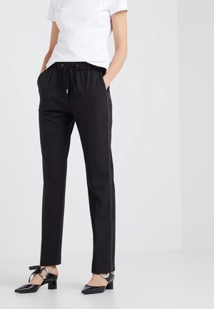 Femme portant un pantalon noir à cordon de serrage et des chaussures noires à bas talon avec des boucles rondes argentées, assortis à un haut blanc à manches courtes.