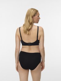 Sieviete ar blondiem matiem, tērpta melnā krūšturī ar regulējamiem siksniņām un melnos augstos apakšbiksēs, redzama no aizmugures uz balta fona.