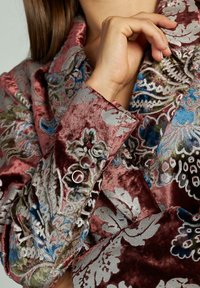Blazer de terciopelo en burdeos con intrincados bordados florales en azul, verde y plata; presenta botones decorativos en el puño.