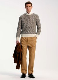 Suéter de punto con un diseño texturizado y estampado en colores oscuros, combinado con pantalones plisados color caqui y zapatos negros, sosteniendo una chaqueta marrón.