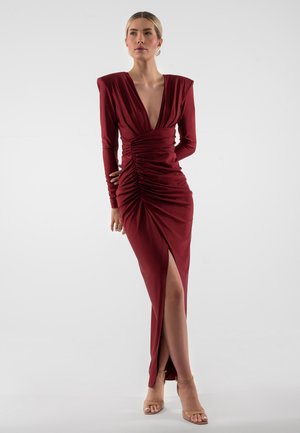 Vrouw draagt een diep rood, geplooid, langemouwen jurk met gewatteerde schouders en een dijhoge split, staand tegen een effen grijze achtergrond.