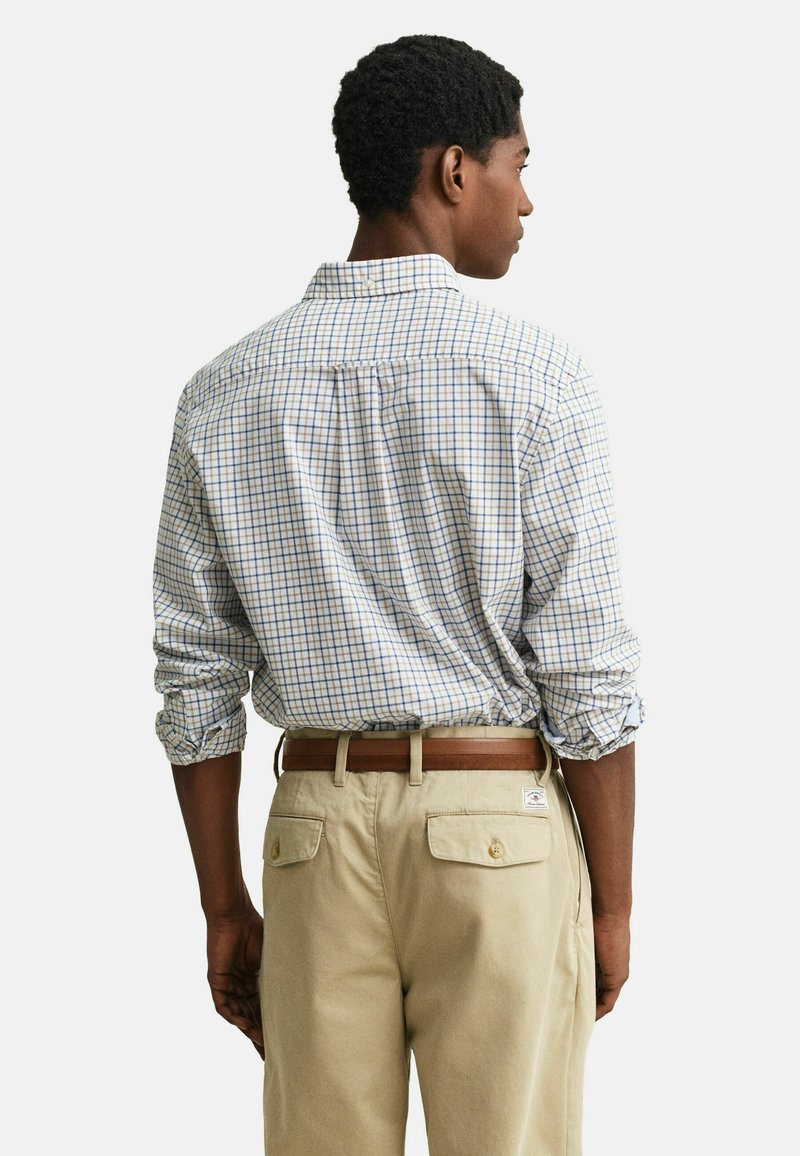 Homme portant un pantalon beige avec des poches arrière et une ceinture marron, associé à une chemise blanche rentrée avec un motif à carreaux bleu et marron.