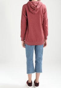 Henkilö, joka on pukeutunut löysään, pitkähiharaiseen pölyruusuisen sävyiseen huppariin, lyhyisiin vaaleansinisiin farkkuihin ja maroonin värisiin lenkkareihin, seisoo valkoista taustaa vasten.