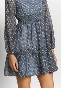 Gråmönstrad klänning med en resårmidja, långa genomskinliga ärmar och en utsvängd kjol. Tyget är lätt med ett rut- och prickmönster.