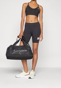 Černý sportovní crop top a šortky, doplněné bílými a černými teniskami. Černá sportovní taška s hladkou texturou a boční kapsou.