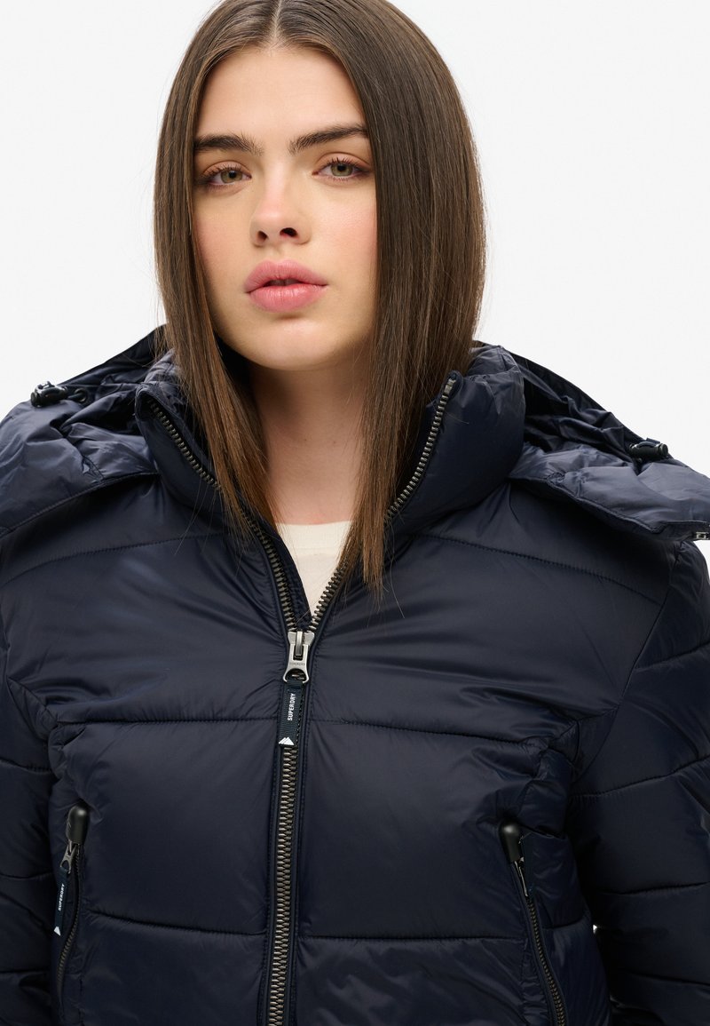 Marineblauwe gewatteerde jas met ritssluiting en capuchon. Heeft een gequilt ontwerp, zijzakken en een gladde, glanzende textuur.