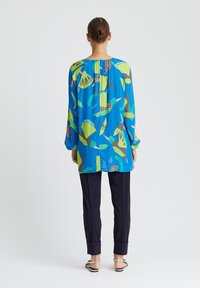 Chemise à manches longues bleu vif avec un motif abstrait coloré featuring des oiseaux et des feuilles ; associée à un pantalon noir ajusté et des sandales plates.