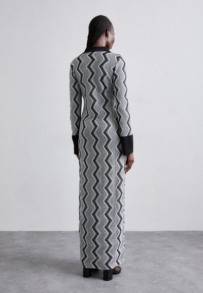 Ilga, figūrą prisitaikanti suknelė su juodai baltu zigzago raštu, pagaminta iš tekstūruotos medžiagos su juodais rankogaliais ir dėvima su masyviomis kulniukais.