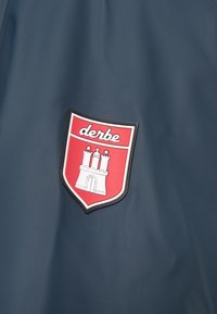 Námořnické svrchní oblečení s obdélníkovou červenou a bílou nášivkou ve tvaru štítu, na které je vyobrazen zámek a slovo "derbe" v bílém písmu.