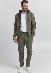 Olive green hoodie and matching joggers with navy side stripes, featuring a logo on the chest and orange accents. White sneakers complete the look.