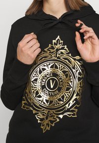 Felpa nera con cappuccio caratterizzata da un grande design grafico dorato con motivi intricati, inclusa un logo circolare che riporta la scritta "VERSACE JEANS COUTURE."