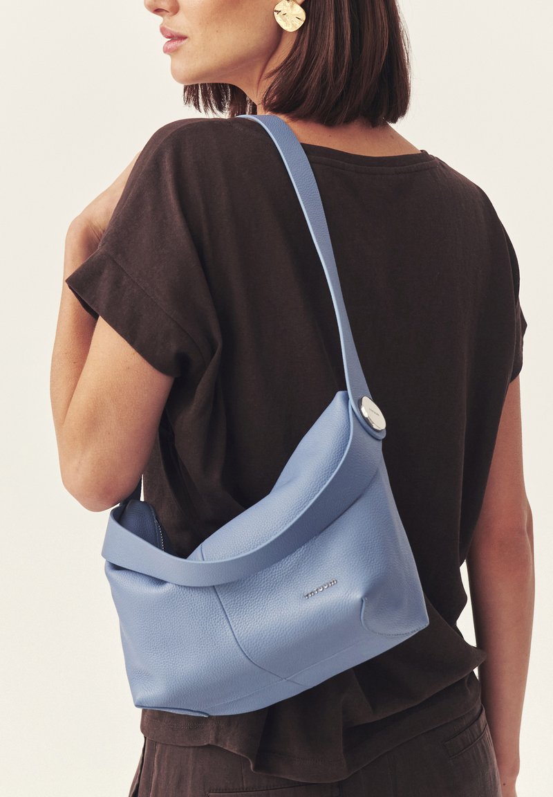 Femme aux cheveux courts et foncés portant des boucles d'oreilles circulaires dorées et un haut noir ample, portant un sac à bandoulière bleu clair texturé.