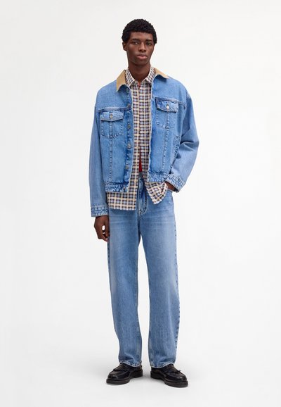 Jeune homme debout portant une veste en jean bleu clair et un jean avec un col beige et une chemise à carreaux, chaussures noires sur fond blanc.